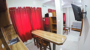 A compact kitchen with a dining area, a red curtain, a flat-screen TV mounted on the wall, and a sofa in the living area.