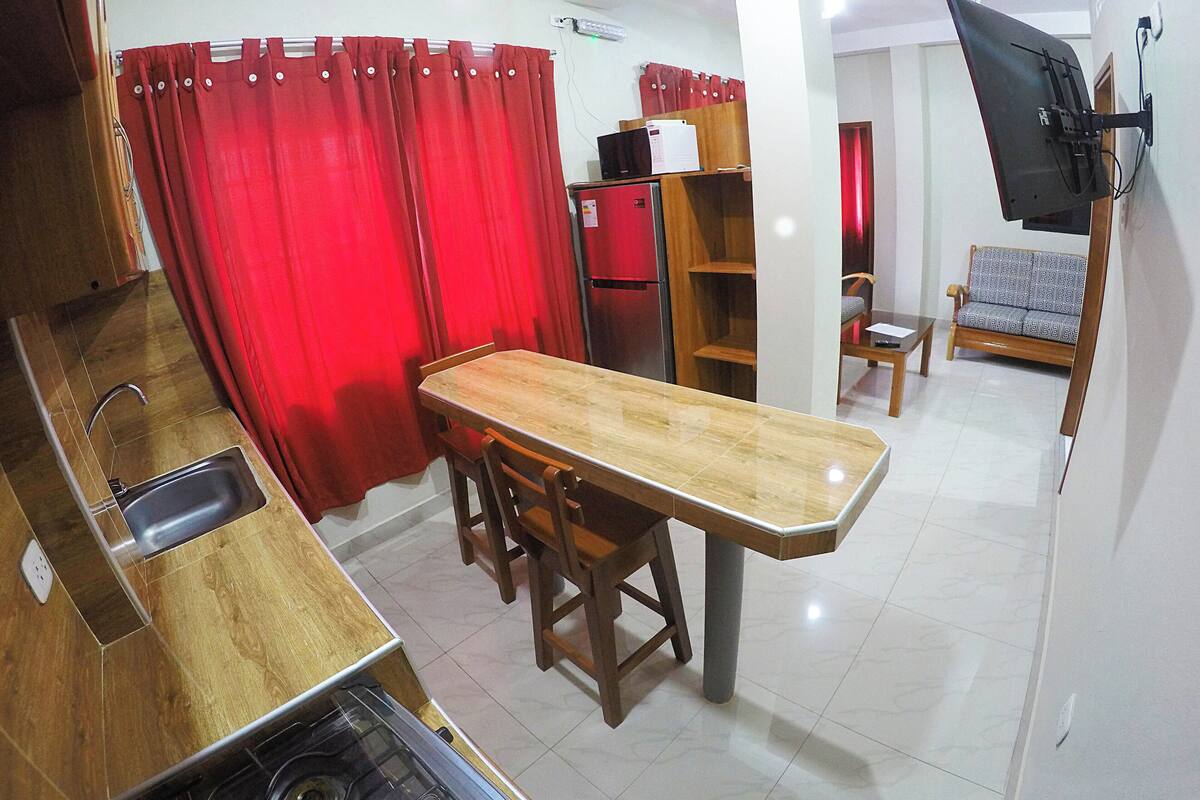 A compact kitchen with a dining area, a red curtain, a flat-screen TV mounted on the wall, and a sofa in the living area.