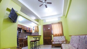 A living room with a sofa, a TV mounted on the wall, a ceiling fan, and a kitchen area with cabinets and a refrigerator.