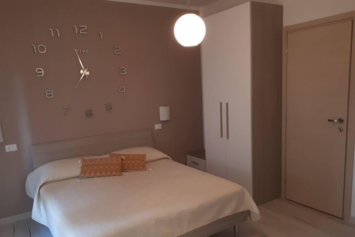 A bedroom with a bed, a wall clock, a wardrobe, and a door.
