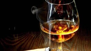 A glass of amber-colored liquid, possibly whiskey, next to a cigar on a wooden surface. - Tahabi Shoe Store (Wazirpur)