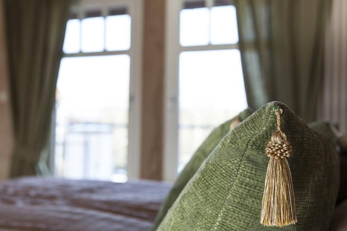 Close-up de um travesseiro verde com um franjado, parte de uma cama em um quarto com janelas amplas e cortinas.