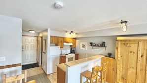 A modern kitchen with wooden cabinets, a white refrigerator, and a built-in stove. There is a wooden dining table with chairs and a small island with bar stools. - Newly Renovated Ground Floor Creekside Studio Condo - Pet Friendly too!  (Silver Star Mountain)