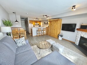 Un salon moderne avec un canapé gris, une table à manger en bois et un coin cuisine doté de meubles en bois et d’un téléviseur fixé au mur.