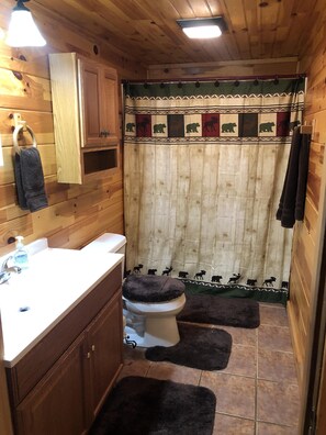 A wooden interior with a white sink, a toilet with a dark rug, and a shower curtain with animal prints. - The AMAZING VIEW at Pop’s Cottage (Bryson City)