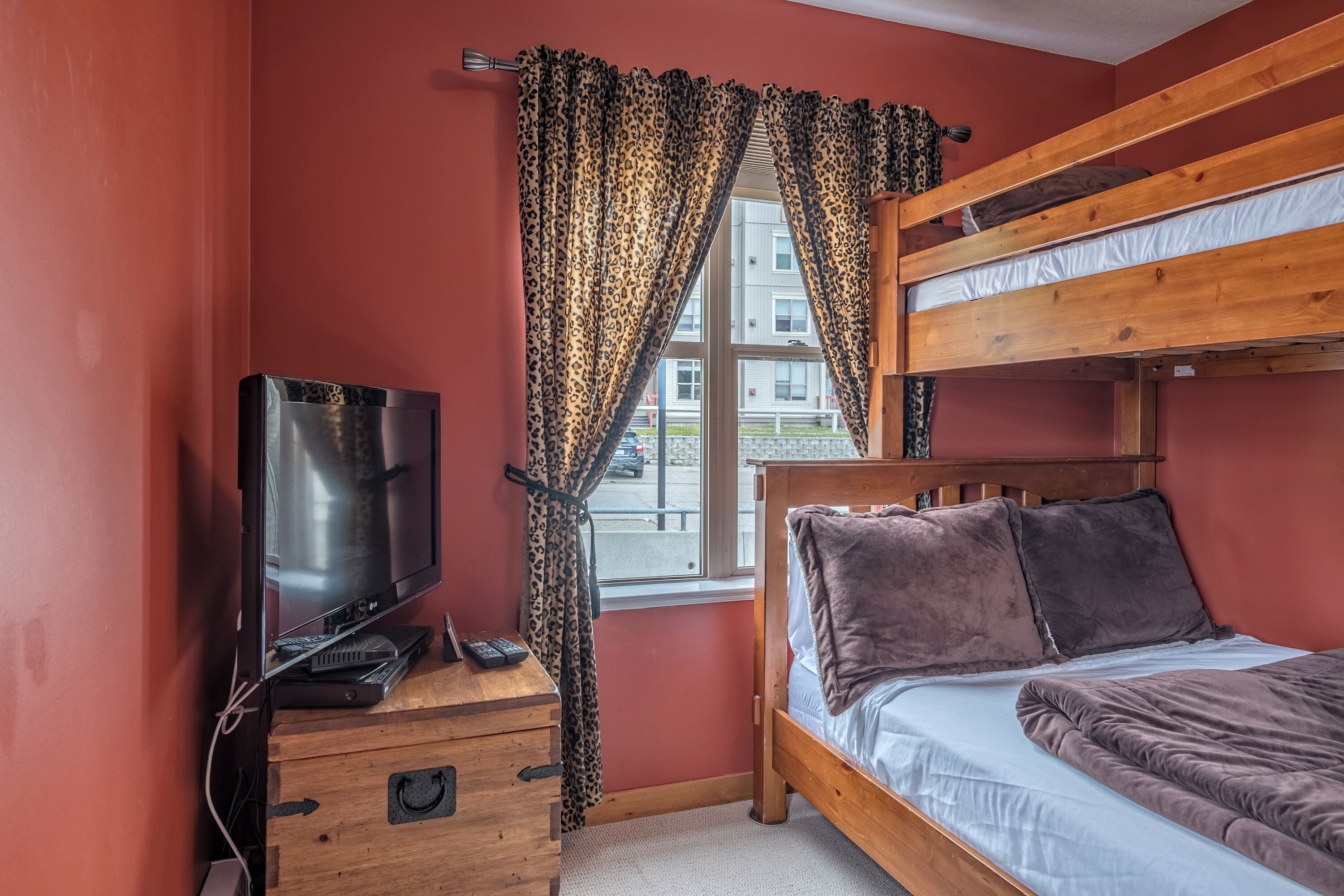 A room with a bunk bed, a television on a wooden dresser, and a window with patterned curtains.