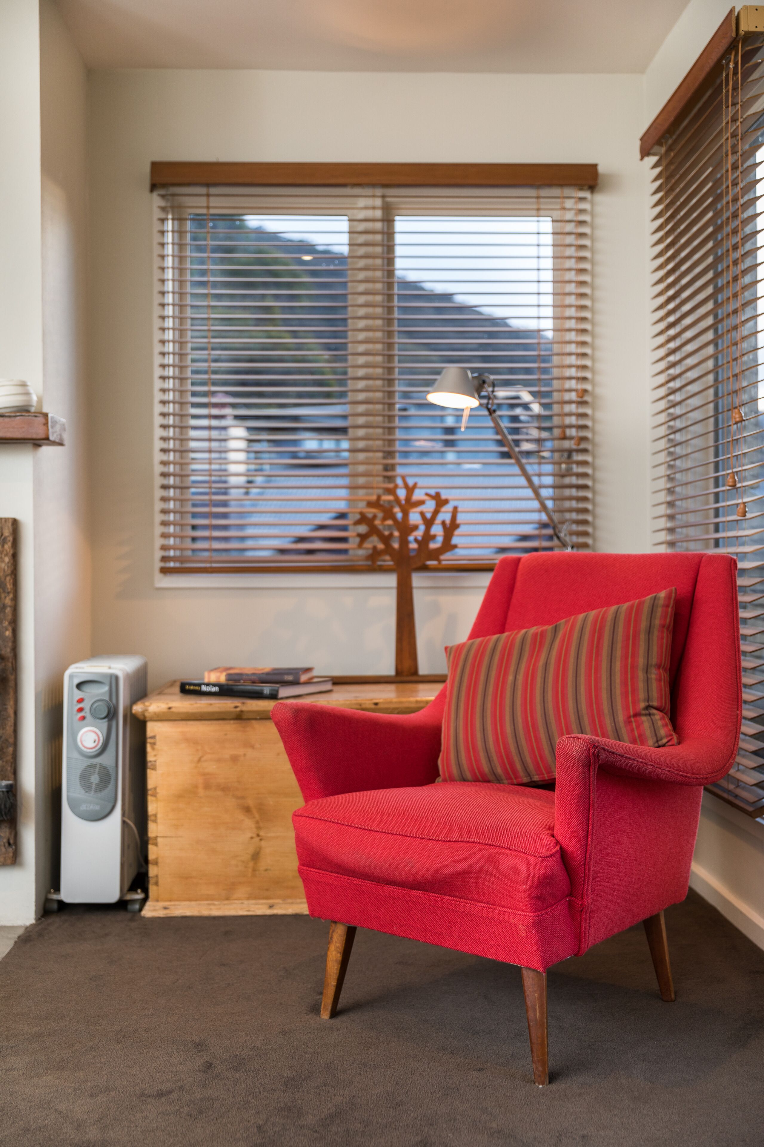A red armchair with a striped pillow, a wooden side table with books, a lamp, and a radiator in a room with a window and blinds.
