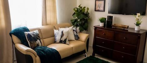 A living room with a beige sofa, a green rug, a wooden dresser, and a large plant.