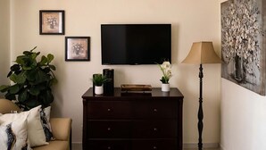 A living room with a dark wooden cabinet, a flat-screen TV, a floor lamp, a sofa with cushions, and a large potted plant. - Walk to Beach, Restaurants  & Attractions (Nassau)