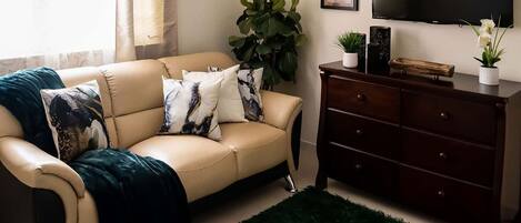 A living room with a beige sofa, a dark wooden dresser, a green rug, and a large plant.