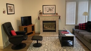 A living room with a fireplace, a television on a wooden stand, a black leather chair, a red pillow, a gray patterned rug, and a sofa with a red and black pillow.