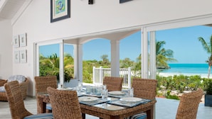 A dining area with wicker chairs and a glass table, overlooking a beach with palm trees and clear blue water. - Coconut Beach Villa! BEACHFRONT Sapodilla! Private Pool! Luxury Family Friendly! (Providenciales)