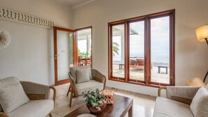 A spacious living room with a large window offering a view of the beach and ocean. There is a comfortable sofa, a wooden coffee table with a floral centerpiece, and a lamp on the right.