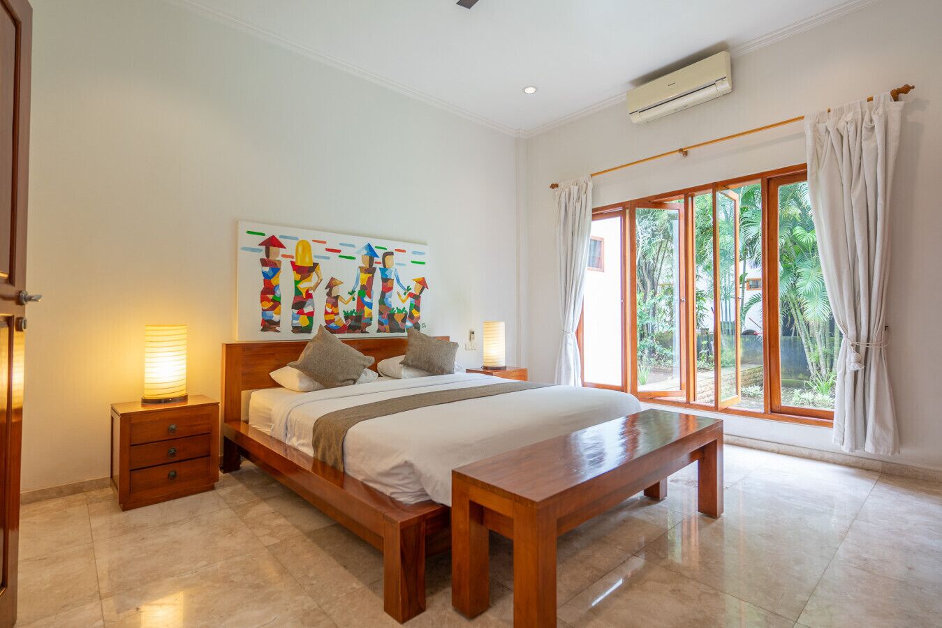 A spacious bedroom with a large bed, wooden nightstands, a bench, and a view of lush greenery through large windows.