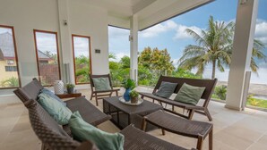 A covered outdoor seating area with wicker furniture and a view of palm trees and a beach.