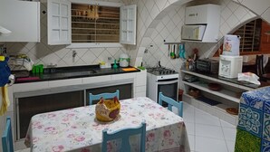 A kitchen with white cabinets, a gas stove, a microwave, and a sink. There is a table with a floral tablecloth and chairs. - Casa em Porto de Galinhas. 450m aproximado da praia. Piscina com apoio coberto (Porto de Galinhas)