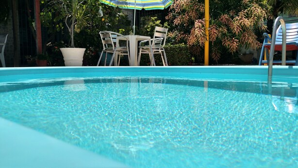 A clear swimming pool with a table and chairs under an umbrella, surrounded by greenery. - Casa em Porto de Galinhas. 450m aproximado da praia. Piscina com apoio coberto (Porto de Galinhas)
