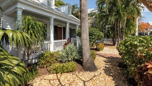 Une maison blanche dotée d’un porche, entourée de plantes tropicales et bordée d’un chemin de gravier.