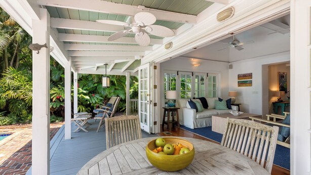 Un espace extérieur abrité comprenant une table à manger, des chaises et un saladier rempli de fruits. On y trouve également une terrasse avec une piscine et une végétation luxuriante en arrière-plan.
