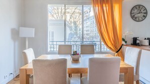 Un comedor con una mesa de madera y cuatro sillas, una ventana con vistas a edificios, un reloj en la pared y una cesta de fruta sobre la mesa.