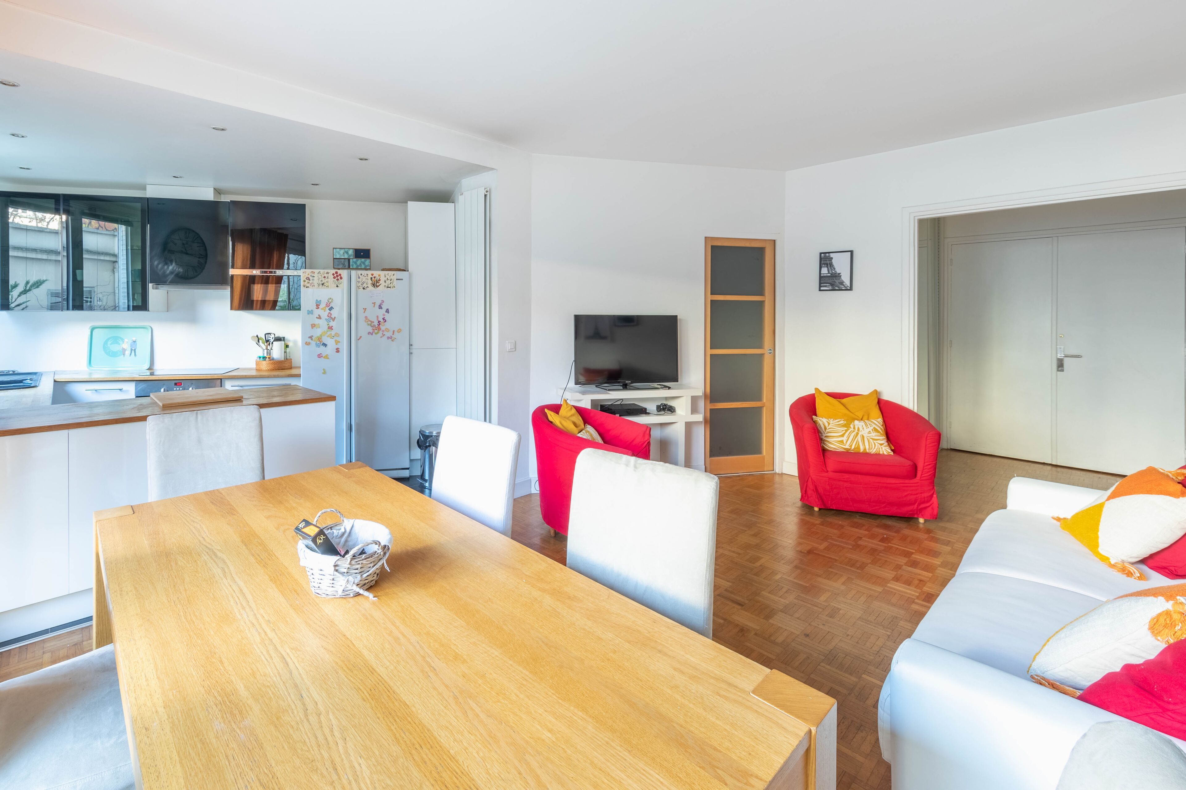 A modern living room with a wooden dining table, white sofas, and a red armchair. A TV is mounted on the wall, and a kitchen area is visible in the background.