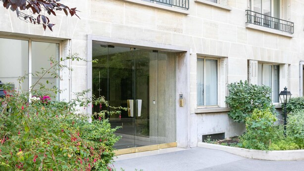 Entrada de un edificio con puerta de cristal, un pequeño jardín con plantas y fachada de piedra.