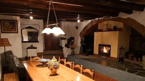Un coin repas chaleureux avec une table et des chaises en bois, un luminaire suspendu et une cheminée en pierre où brûle un feu.