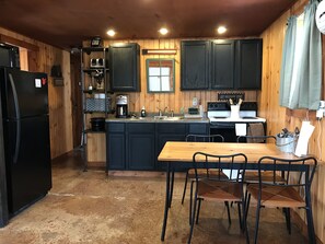 A compact kitchen with a refrigerator, stove, sink, and dining area. - Cozy Fireside Cabin, Great for Couples, Families and Outdoorsmen! (McArthur)