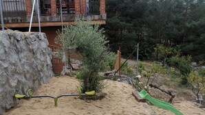 A sandy play area with a slide and a small tree, a stone wall, and a building with a balcony in the background. - Los Corzos Burgohondo Avila (Burgohondo)