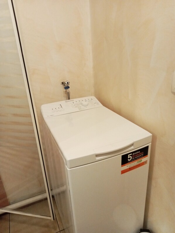 A compact, white washing machine in a bathroom with a tiled floor and a glass shower door.