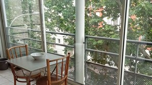 Un comedor con mesa y sillas, ventanas amplias con vistas a árboles y edificios, y barandilla de balcón.