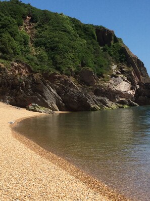 A pebble beach with clear water, a rocky cliff, and dense greenery.