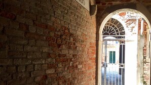 A brick corridor with a sign indicating a historical site, leading to an open area with a metal gate and a view of a building with a green door. - Ca' Venexiana, quiet 2 bedroom with private canalside courtyard - no added fees! (Venezia)