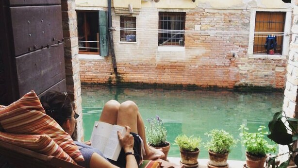 A person relaxing on a wooden chair by a brick wall with a small pool, surrounded by potted plants. - Ca' Venexiana, quiet 2 bedroom with private canalside courtyard - no added fees! (Venezia)