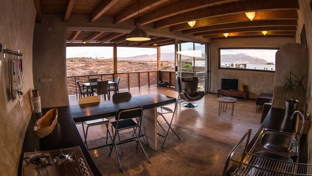 Amplia sala de estar con techo de madera, mesa de comedor y sillas. Una ventana grande ofrece vistas a las montañas.