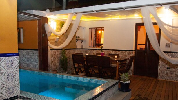 Una piscina con borde de azulejos, un área de comedor con sillas y una mesa, y una puerta que da a otra habitación.