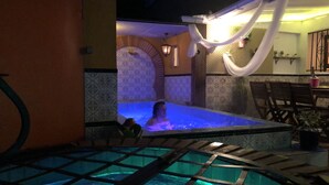 Zona de piscina con una persona en el agua, rodeada de paredes con diseños y una zona de estar cubierta.