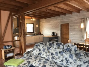 A cozy living room with a patterned sofa, wooden beams, a kitchen with a sink and cabinets, and a dining area with a table and chairs.