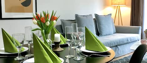 A dining area with a table set for four, a sofa in the background, and a framed picture on the wall.