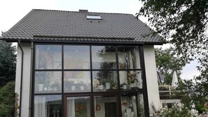 A two-story house with large windows, a tiled roof, and a patio with a table and chairs.
