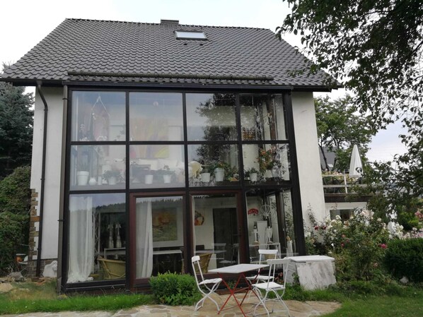 A two-story house with large windows, a tiled roof, and a patio with a table and chairs.