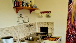 A compact kitchen with a coffee maker, a sink, and a microwave. There are two hanging mugs and a shelf with kitchen items.