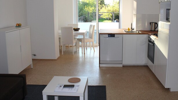 A modern kitchen with white cabinetry, a dishwasher, and a coffee maker. There is a dining area with a table and chairs, and a view of greenery outside. - House Roy, 3 room apartment in the redeveloped house  (Zülpich)