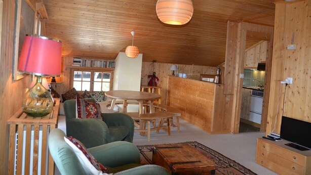 Interior acogedor de una cabaña con comedor, sofá, televisor y cocina visible a través de un arco.