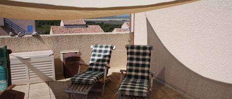 A rooftop terrace with two lounge chairs and a table, a white wall, and a view of buildings in the background.