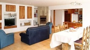 Un salon avec un canapé, un téléviseur et une cheminée, un coin repas avec une table et des chaises, et une cuisine équipée de placards et d’appareils électroménagers.