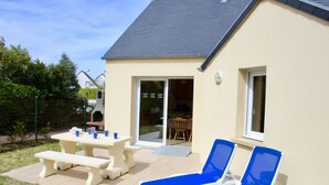 Una casa con patio, dos sillones azules, una mesa de picnic y un ventanal corredizo que da a un espacio interior.