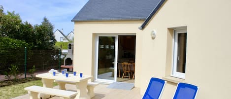 A house with a patio, two blue lounge chairs, a picnic table, and a sliding glass door leading to an indoor area.