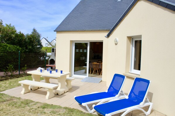 A house with a patio, two blue lounge chairs, a picnic table, and a sliding glass door leading to an indoor area.