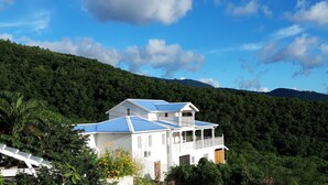 Un edificio blanco con techo azul, rodeado de abundante vegetación y colinas.